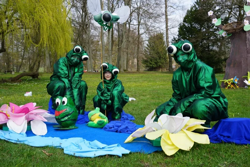 Frösche am Teich im Märchen-Musical – Drei Darsteller als grüne Frösche hocken an einem blauen Teich, umgeben von übergroßen Blüten und Requisiten, über ihnen schwebt ein Insektenstab. Fröhliche Frühlingsszene im Park aus Abenteuer mit dem Hasensohn.