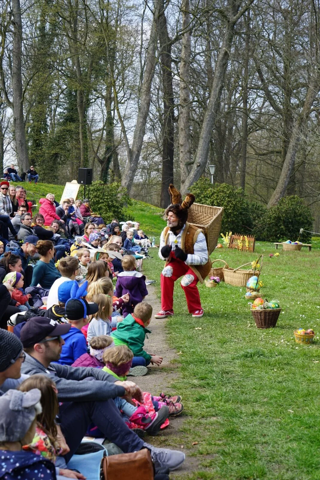 Osterhase begrüßt Publikum mit Kindern im Park – Der Osterhase mit großem Rückenkorb spricht vor einer langen Reihe sitzender Kinder und Familien im Freien, entlang eines Wegs mit Körben voller bunter Ostereier. Open-Air-Szene aus Abenteuer mit dem Hasensohn.