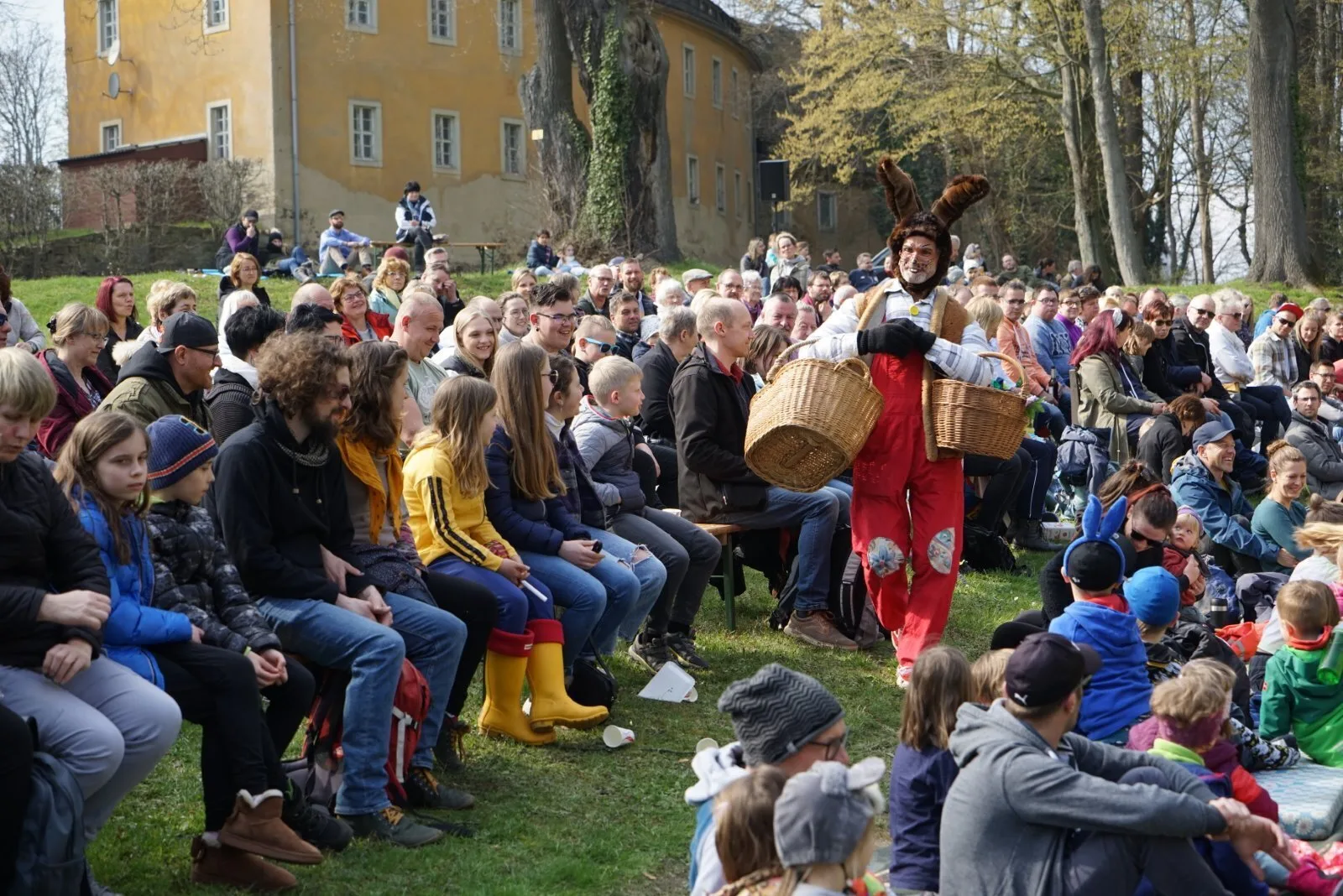 Osterhase mit Körben im vollbesetzten Publikum – Zwischen dicht besetzten Bänken läuft der Osterhase mit zwei großen Weidenkörben vorbei, viele lachende Gesichter auf der Theaterwiese. Fröhliche Szene aus Abenteuer mit dem Hasensohn.