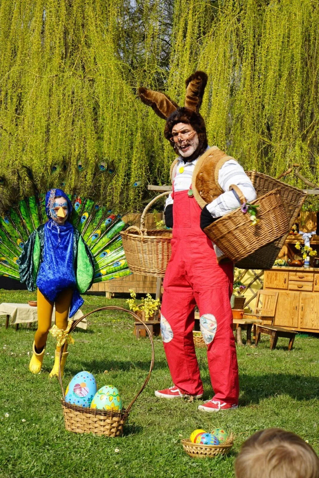 Osterhase und Pfau im Frühlingsgarten – Der Osterhase in roter Latzhose trägt Körbe, neben ihm steht ein prächtiger Pfau in leuchtendem Kostüm mit Federfächer. Vorne liegen bemalte Ostereier in Körben, Bühne im Grünen.