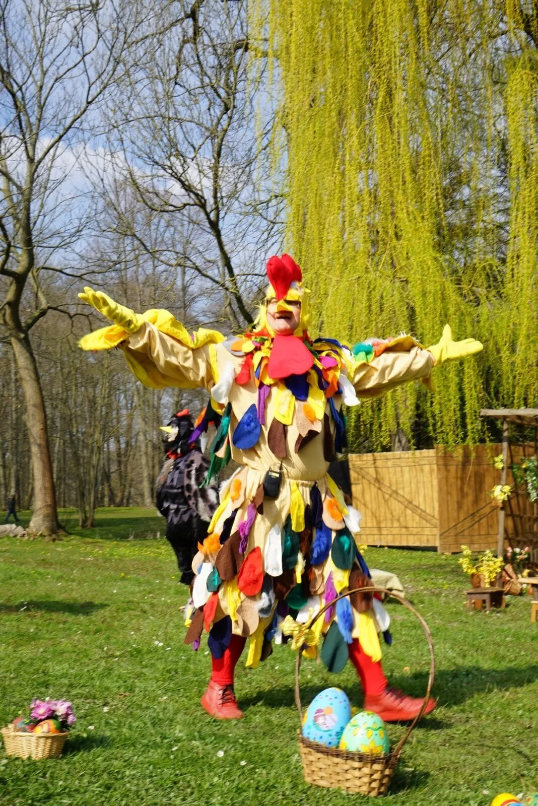 Bunter Hahn auf der Wiese mit Ostereiern – Ein farbenfroher Hahn in großem Federkostüm breitet die Arme aus, neben ihm ein Korb mit bemalten Eiern, hinten zieht eine Hühnerschar vorbei. Heitere Open-Air-Szene im Osterdorf.