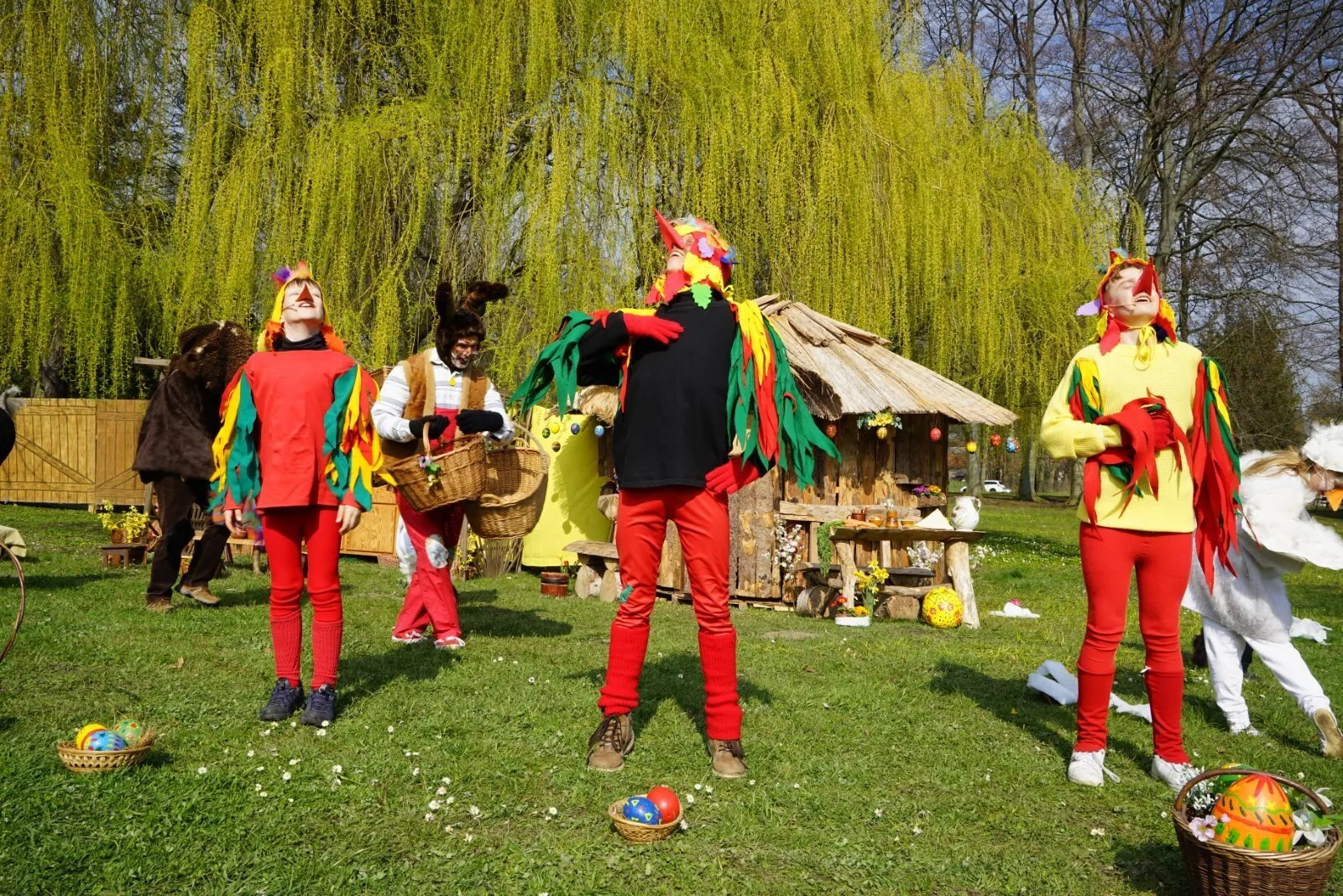 junge Hähne auf der Osterwiese – Drei bunt gefiederte junge Osterhähne vom Bauernhof stehen vor einer rustikalen Holzhütte mit Osterdeko, der Osterhase arbeitet im Hintergrund. Kleine Körbe mit Eiern liegen im Gras, fröhliche Frühlingsstimmung.
