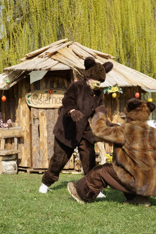 Bären toben vor dem Osterhasenhaus – Zwei Darsteller in braunen Bärenkostümen rangeln auf der Wiese vor einem rustikalen Holzhaus mit Schild Osterhasen, umgeben von bunter Frühlingsdekoration. Humorvolle Szene aus dem Musical.