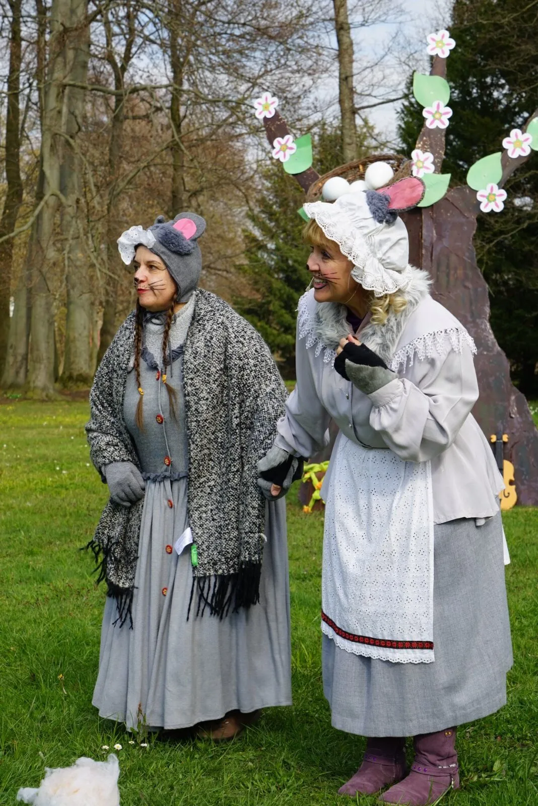 Mäusefrauen schauen nach den Osterhasen – Zwei Darstellerinnen als Mäuse in grauen Kleidern und Hauben stehen lachend auf der Wiese, neben ihnen blühende Requisiten und Frühlingsbäume. Szene aus Abenteuer mit dem Hasensohn im Osterdorf.