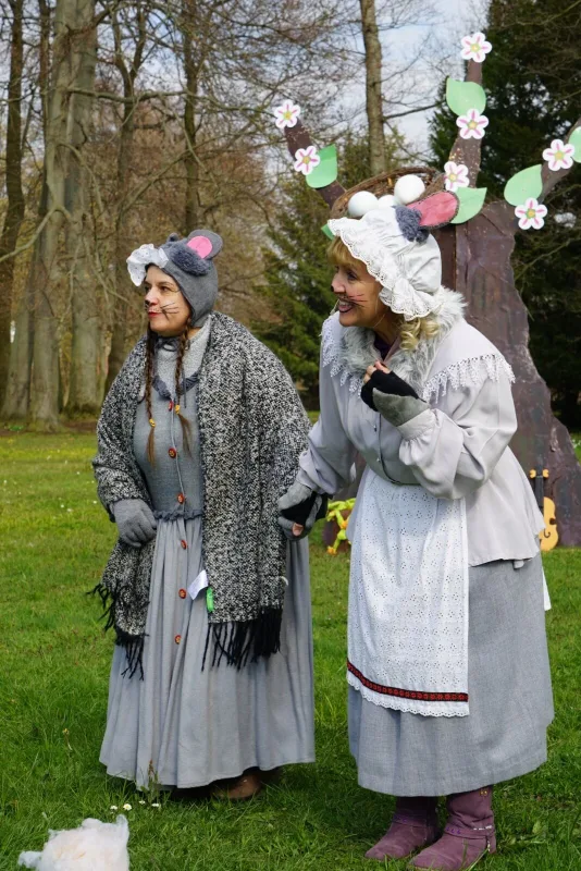 Mäusefrauen schauen nach den Osterhasen – Zwei Darstellerinnen als Mäuse in grauen Kleidern und Hauben stehen lachend auf der Wiese, neben ihnen blühende Requisiten und Frühlingsbäume. Szene aus Abenteuer mit dem Hasensohn im Osterdorf.