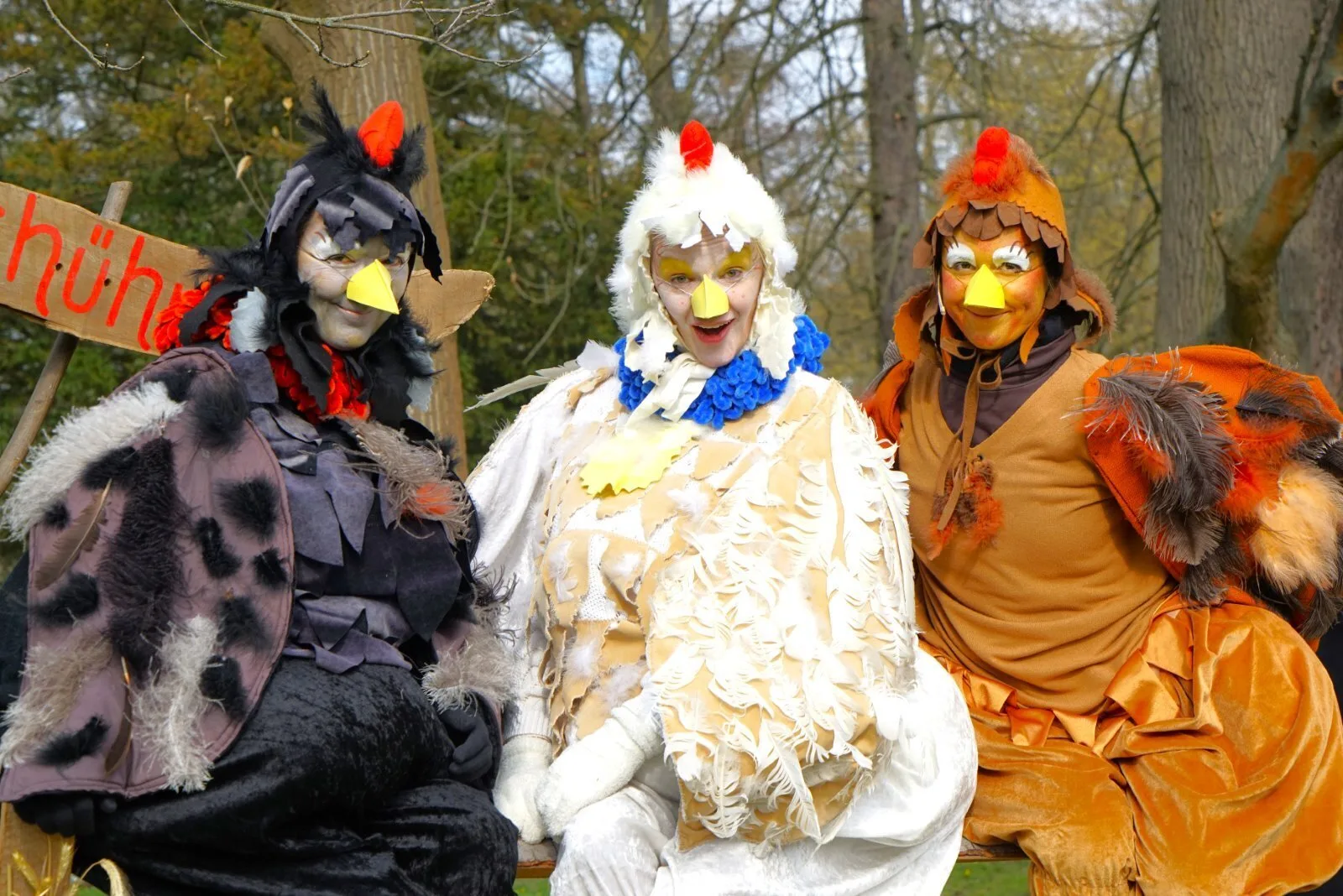 Drei Hennen vom Hühnerhof im Kostüm – Drei Darstellerinnen als Hennen in Schwarz, Weiß und Braun sitzen Arm in Arm vor einem Holzschild, bunte Stofffedern und Schnäbel prägen die humorvolle Gruppe. Porträtszene aus Abenteuer mit dem Hasensohn.