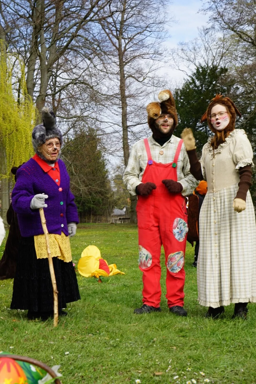 Hasenfamilie mit Großmutter auf der Wiese – Großmutter mit Stock, der Hasensohn Später in roter Latzhose und seine Freunding, die junge Häsin Vorher stehen zusammen auf der Wiese und blicken zum Publikum, Frühling im Osterdorf mit Bäumen und Requisiten.