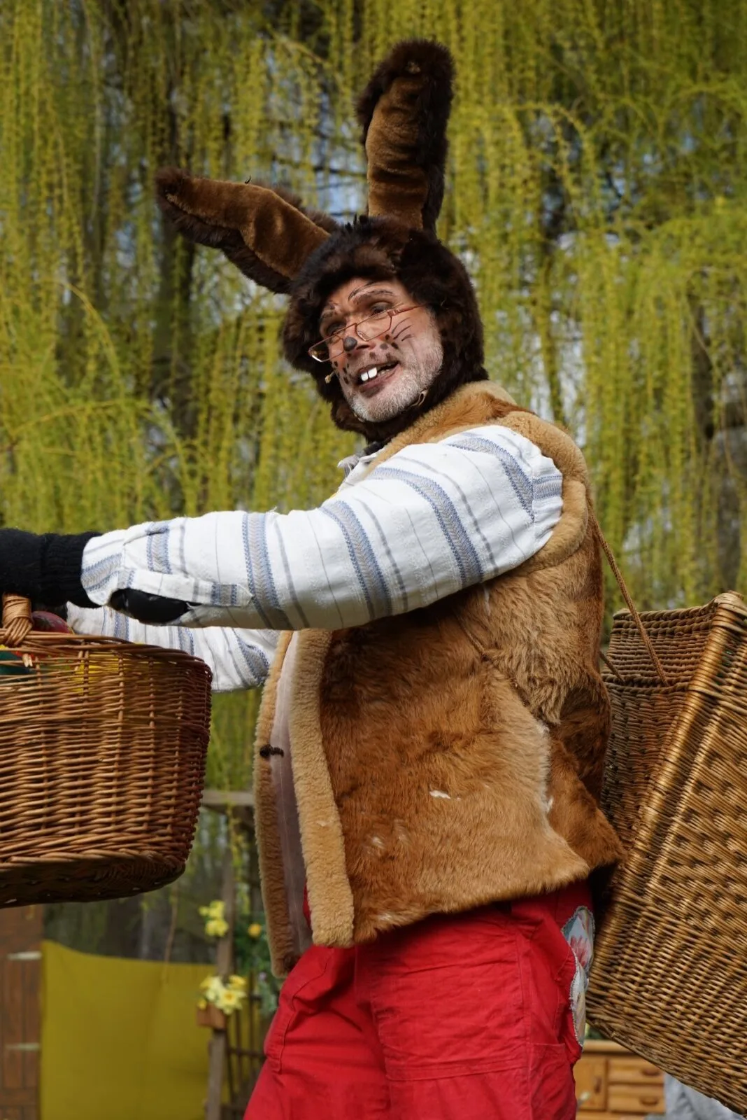 Osterhase mit Körben im Porträt – Der Osterhase in gestreiftem Pullover und Fellweste trägt Weidenkörbe über Schulter und Arm, lächelt mit schiefen Zähnen in die Kamera, Weiden im Hintergrund. Charakterporträt der Hauptfigur.