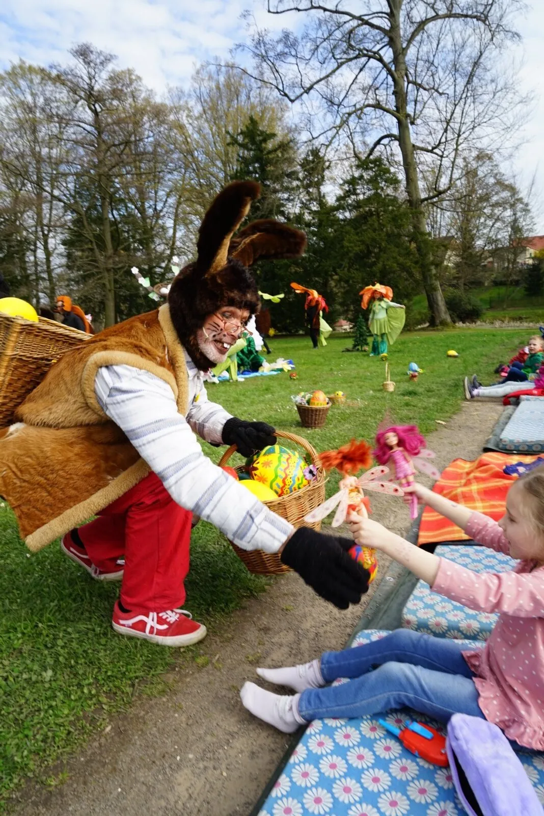 Osterhase schenkt Ei an Kind im Publikum – Der Osterhase kniet am Wegesrand und reicht einem Mädchen auf bunten Matten ein bemaltes Ei, im Korb liegen weitere Ostereier, hinter ihm Wirbel aus Tanz und Kostümen. Herzerwärmende Publikumsnähe.