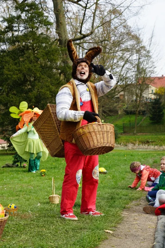 Osterhase ruft dem Kinderpublikum zu – Der Osterhase mit zwei großen Weidenkörben ruft den Kindern am Wegesrand zu, im Hintergrund tanzen Blütenwesen auf der Wiese. Fröhliche Interaktion beim Frühlingsfest von Abenteuer mit dem Hasensohn.
