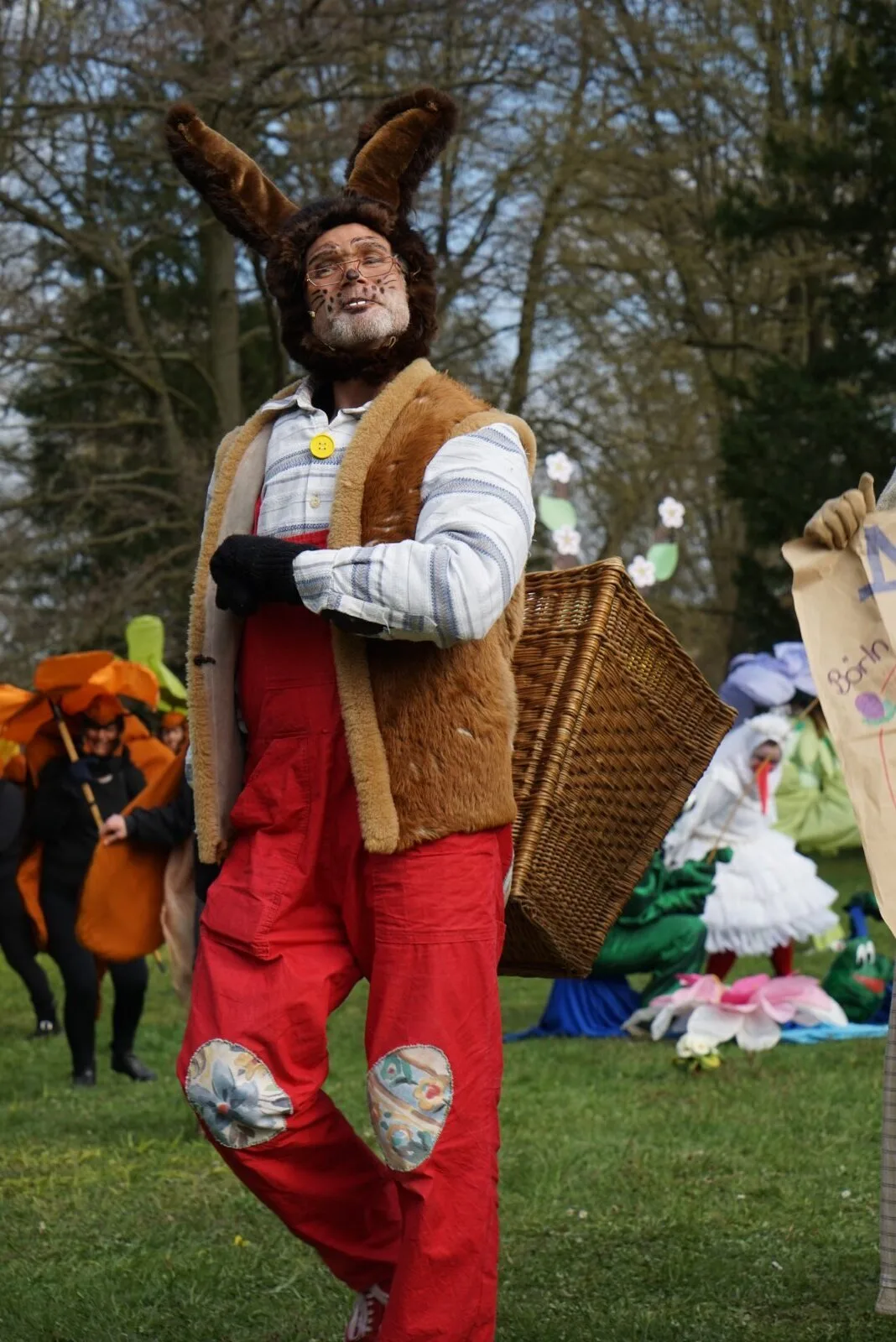Osterhase mit Rückenkorb auf der Wiese – Der Osterhase schreitet mit großem Rückenkorb an Frühlingsfiguren vorbei und singt, rundherum blühen die Wiesen und Requisiten. Dynamische Szene der Open Air Aufführung Abenteuer mit dem Hasensohn.