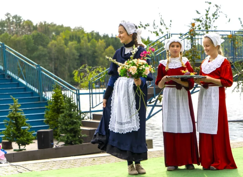 Bäuerinnen mit Blumenstrauß und Tablett am Seeufer – Drei Bäuerinnen in Tracht mit weißen Hauben stehen vor der blauen Brücke am See, eine hält einen Blumenstrauß, zwei tragen ein Tablett mit Häppchen, ländliche Szene zwischen Markt und Spazierweg.