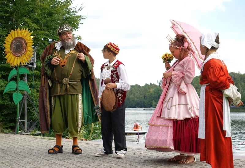 König, Prinzessin und Diener am Seeufer – Der König im Jagdgewand spricht mit dem Diener, die Prinzessin in Rosa mit Sonnenschirm hört zu, eine Zofe steht neben ihnen