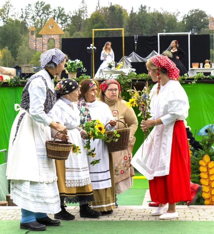 Bäuerinnen mit Körben und Blumen beraten sich am Dorfmarkt – Bäuerinnen in Tracht stehen mit Körben und Sonnenblumen zusammen und beraten sich vor grüner Bühnenwand, oben agieren zwei Geister auf der Galerie – ländliche Marktszene mit Blumen.