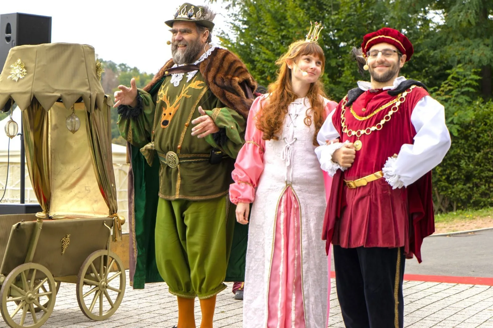 König, Prinzessin und Graf Carabas neben der goldenen Kutsche – Der König im Jagdgrün präsentiert lächelnd die Prinzessin und den jungen Grafen Carabas neben einer goldenen Kutsche, feierlicher Moment der Annäherung nach der Rettung am Seeufer.
