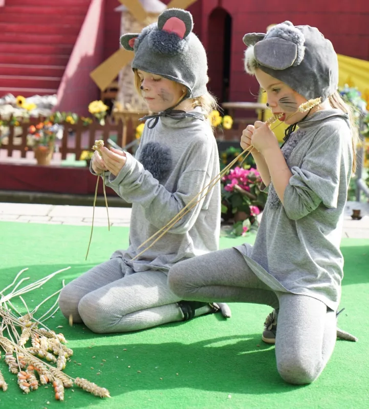 Mäusekinder knabbern Ähren auf der grünen Bühne – Zwei als Mäuse verkleidete Kinder knien auf der grünen Bühne und knabbern an Ähren, im Hintergrund rote Treppen und Blumen – Dorffest rund um Feld, Mühle und Ernte.