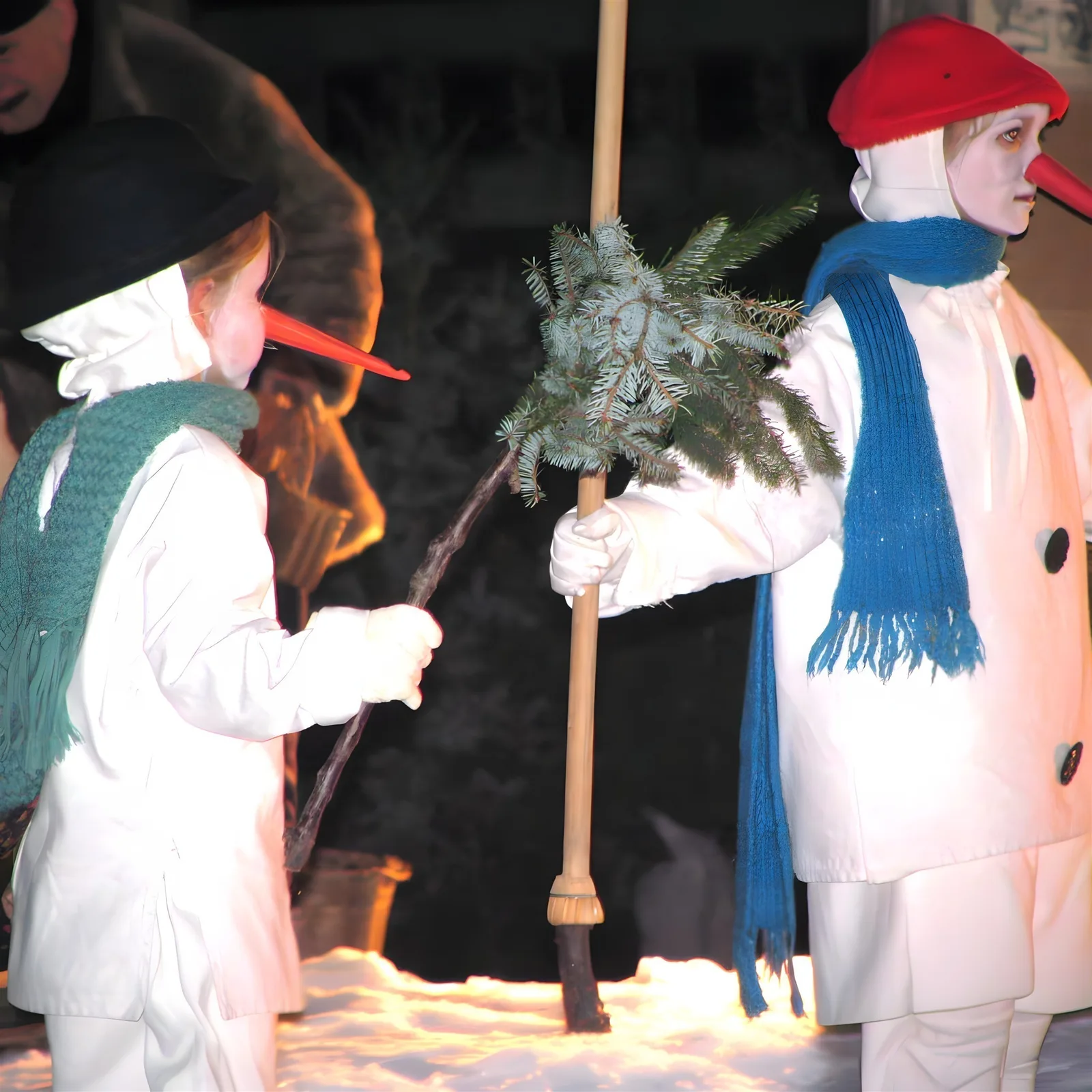 Schneemannkinder im Winterzauber ? Zwei verkleidete Schneemannkinder mit Möhrennasen und bunten Mützen stehen im Bühnen-Schnee, sie tragen Schals und halten Besen sowie Zweigbündel – humorvolle Eröffnung der frostigen Reise.