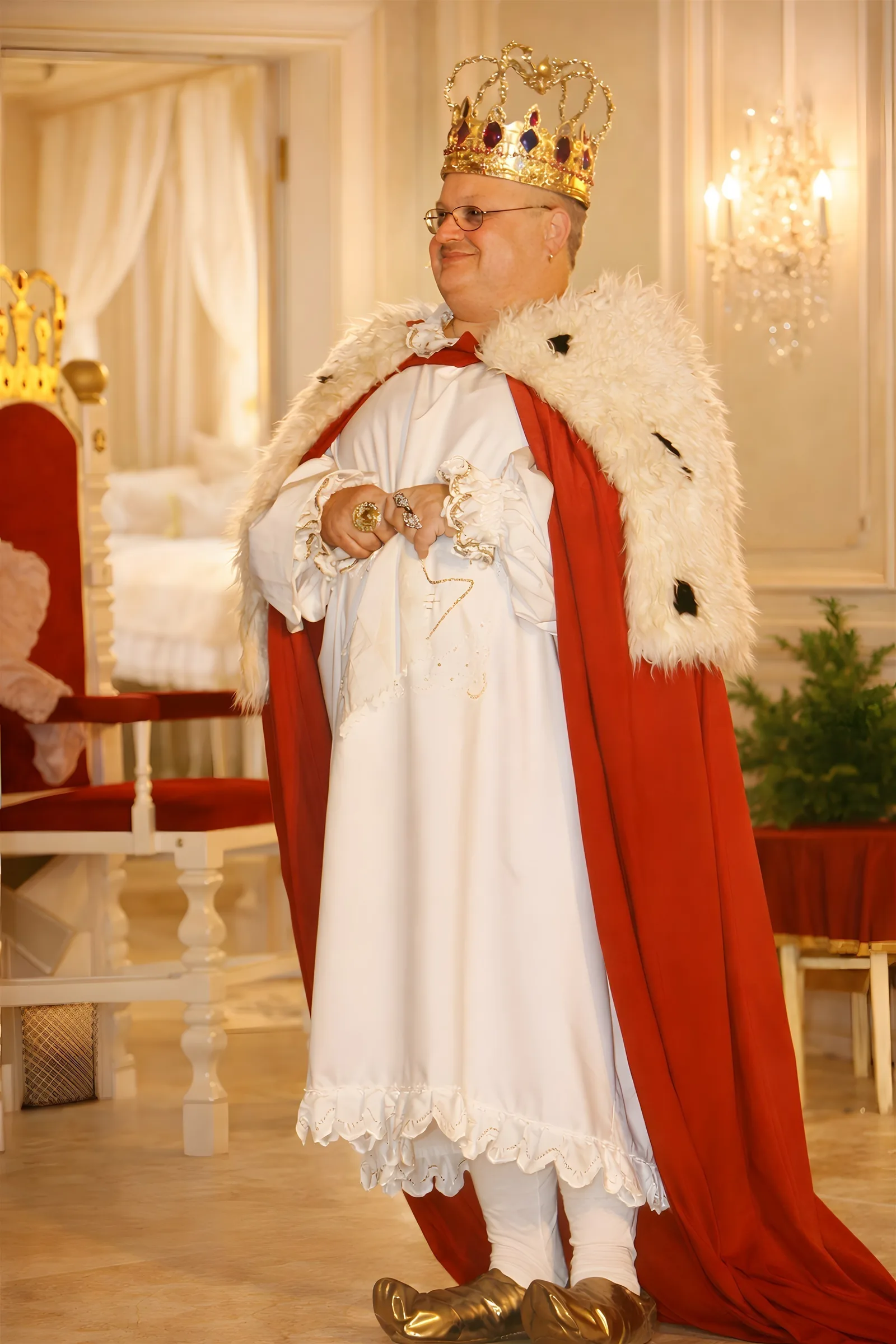 König in rotem Hermelinmantel ? Der König sitzt lächelnd mit goldener Krone und rotem Hermelinmantel im hellen Thronsaal, Kerzen und weiße Vorhänge rahmen die Szene – warmes Königsporträt.