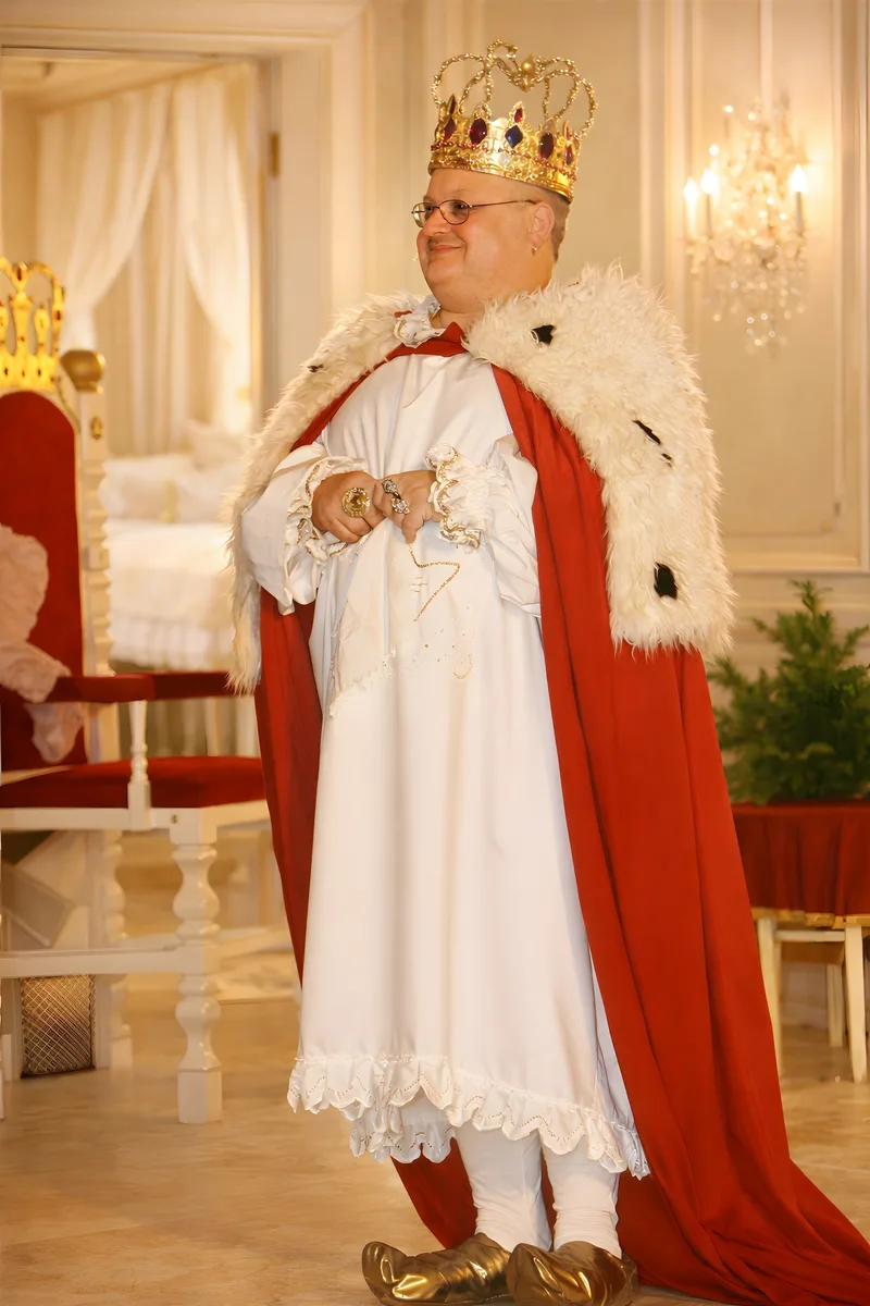 König in rotem Hermelinmantel ? Der König sitzt lächelnd mit goldener Krone und rotem Hermelinmantel im hellen Thronsaal, Kerzen und weiße Vorhänge rahmen die Szene – warmes Königsporträt.