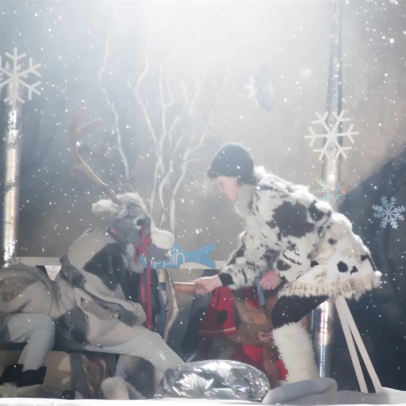 Rentier wird im Schneesturm versorgt ? Im tanzenden Schnee füttert eine Pelzfigur das erschöpfte Rentier, weiße Bäume und Flocken umgeben die Bühne – Fürsorge mitten im Winter.