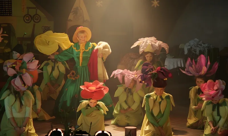 Blumenwiese der Zauberfrau ? Eine Zauberfrau in grünem Kleid mit Sonnenhut steht zwischen Kinderblumen in leuchtenden Kostümen – verzauberte Gartenwelt.