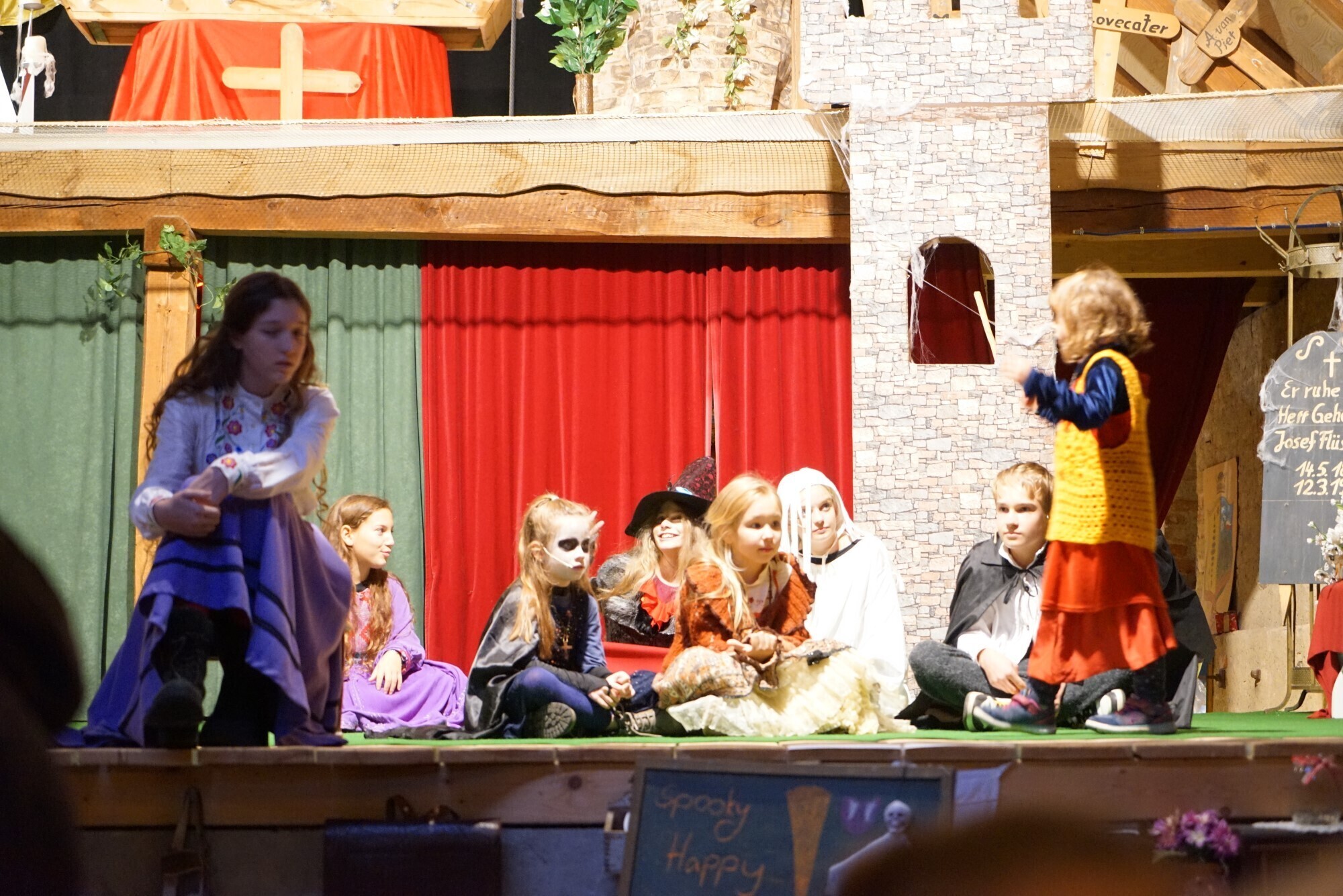 Kinder auf der Bühne beim Halloween-Kürbisfest. Kinder in bunten Kostümen spielen Szenen des Märchen-Musicals „Kürbisfest und Maskenball mit Graf Dracula“ auf der Bühne, mit Vampir- und Hexenmasken vor Burgkulisse.