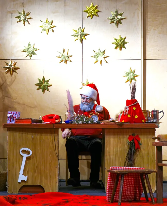 Weihnachtsmann im himmlischen Büro unter Sternen – Der Weihnachtsmann sitzt an seinem Holztisch mit roter Mütze, Gänsekiel und Telefon, über ihm glänzen goldene Sterne – emsige Vorbereitung auf Geschenke und Wunschlisten im Weihnachtsbüro.