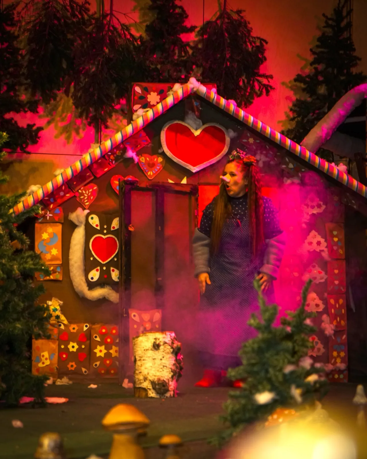 Trude am Knusperhäuschen im dunklen Wald – Vor einem bunt verzierten Lebkuchenhaus mit Herzen tritt die Hexe aus der Tür, Nebel und Tannen umrahmen die Szene – Anspielung auf das Knusperhaus aus Hänsel und Gretel.