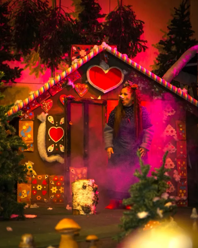 Trude am Knusperhäuschen im dunklen Wald – Vor einem bunt verzierten Lebkuchenhaus mit Herzen tritt die Hexe aus der Tür, Nebel und Tannen umrahmen die Szene – Anspielung auf das Knusperhaus aus Hänsel und Gretel.