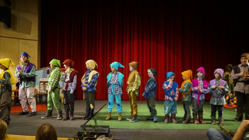 Wichtel in Reihe – Eine lange Reihe bunt gekleideter Wichtel mit Zipfelmützen steht auf der Bühne, bereit für die Werkstatt des Weihnachtsmanns – fröhliche Helfer in der Weihnachtsgeschichte.