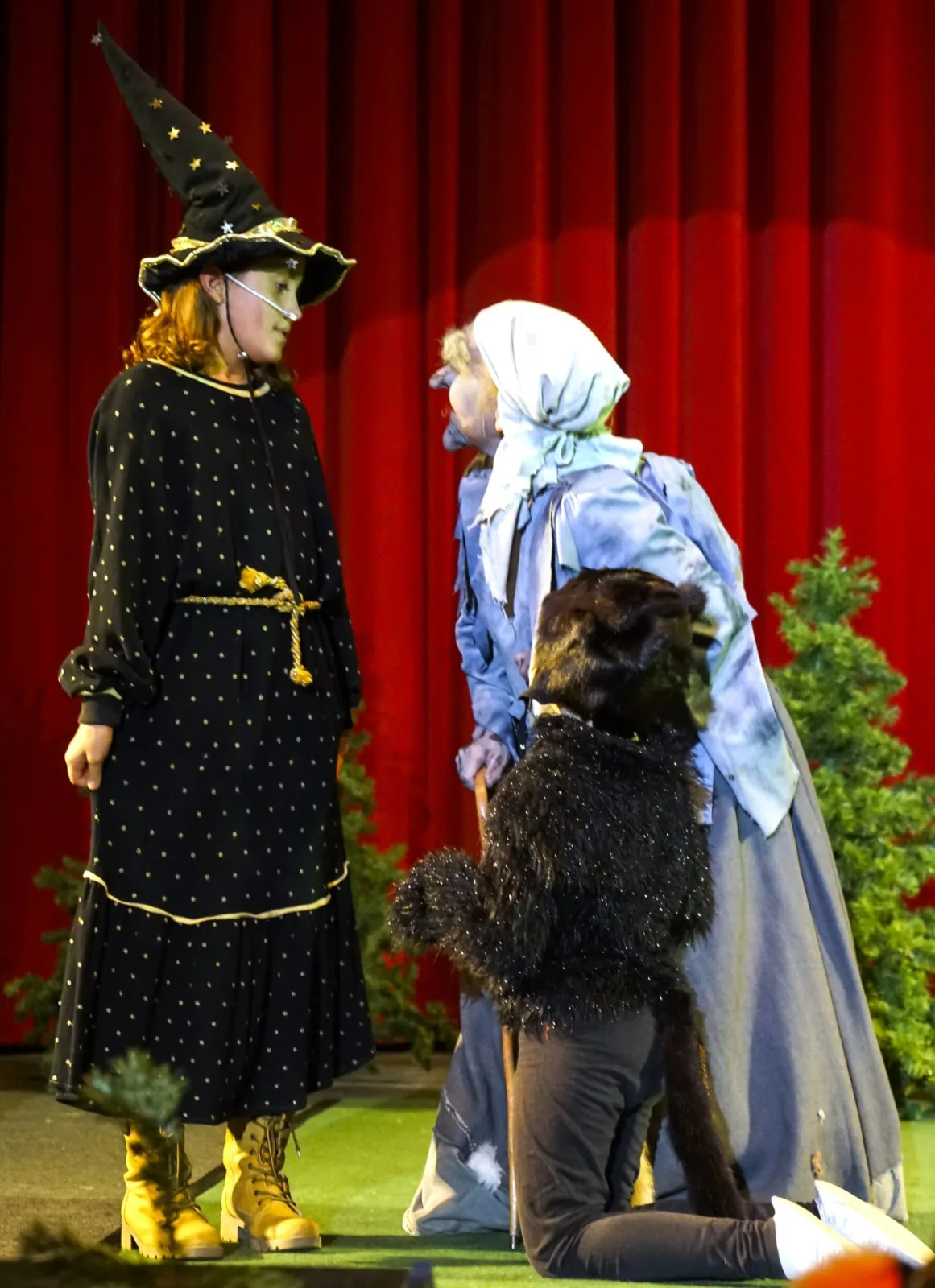 junge Hexe im Gespräch mit der Knusperhexe und einer schwarzen Katze – Eine junge Hexe im schwarzen Sternenkleid spricht mit der Knusperhexe, daneben kniet ein als schwarze Katze verkleidetes Kind – geheimnisvolle Waldbegegnung vor rotem Vorhang.