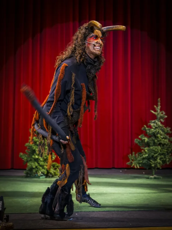 Teufelchen mit Hörnern beim Auftritt – Teufelkind in dunklem Gewand mit Hörnern schreitet über die Bühne, hinter ihm Tannen und roter Vorhang – kraftvoller Auftritt der düsteren Figur.