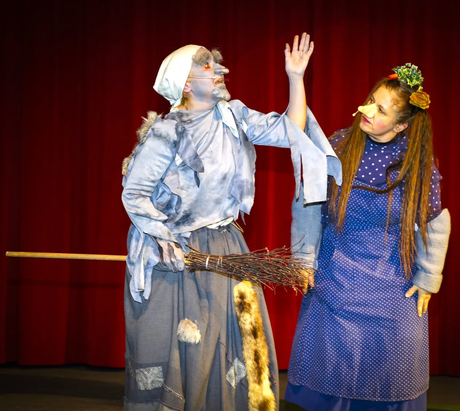 Alte Knusperhexe mit Besen und Trude im Dialog – Eine grau geschminkte alte Frau mit Kopftuch und Reisigbesen hebt warnend die Hand, neben ihr steht die Trude mit langen Zöpfen und Blätterkranz vor rotem Vorhang – gespannte Waldszene.