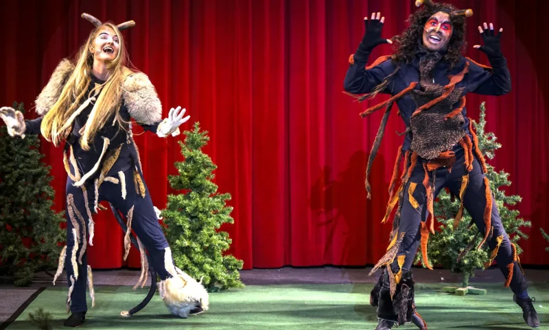 Teufelkinder tanzen mit Hörnern – Zwei Teufelkinder in zottigen Kostümen tanzen vor rotem Vorhang zwischen Tannen, wildes Spiel und starke Mimik bringen Schwung in die Szene.