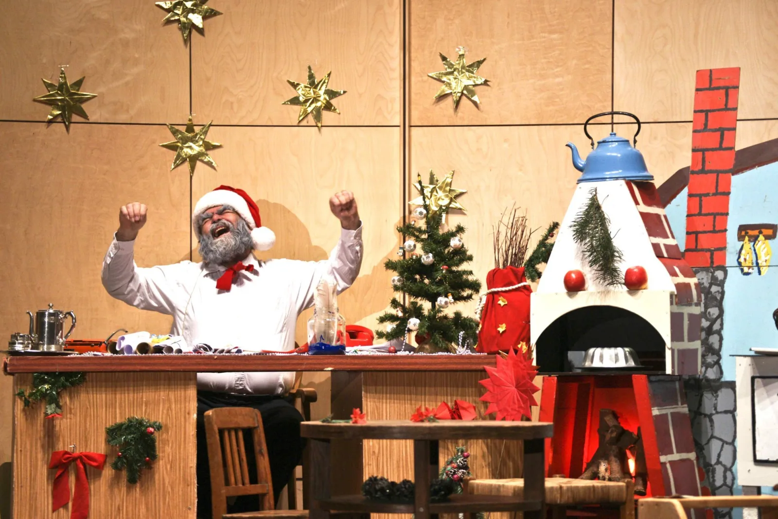 Weihnachtsmann gähnt im Weihnachtsbüro – Der Weihnachtsmann reißt vor Freude die Arme hoch an seinem Schreibtisch mit Christbaum, Kaminattrappe und Sternen an der Wand – siegreicher Moment im Geschenkechaos.