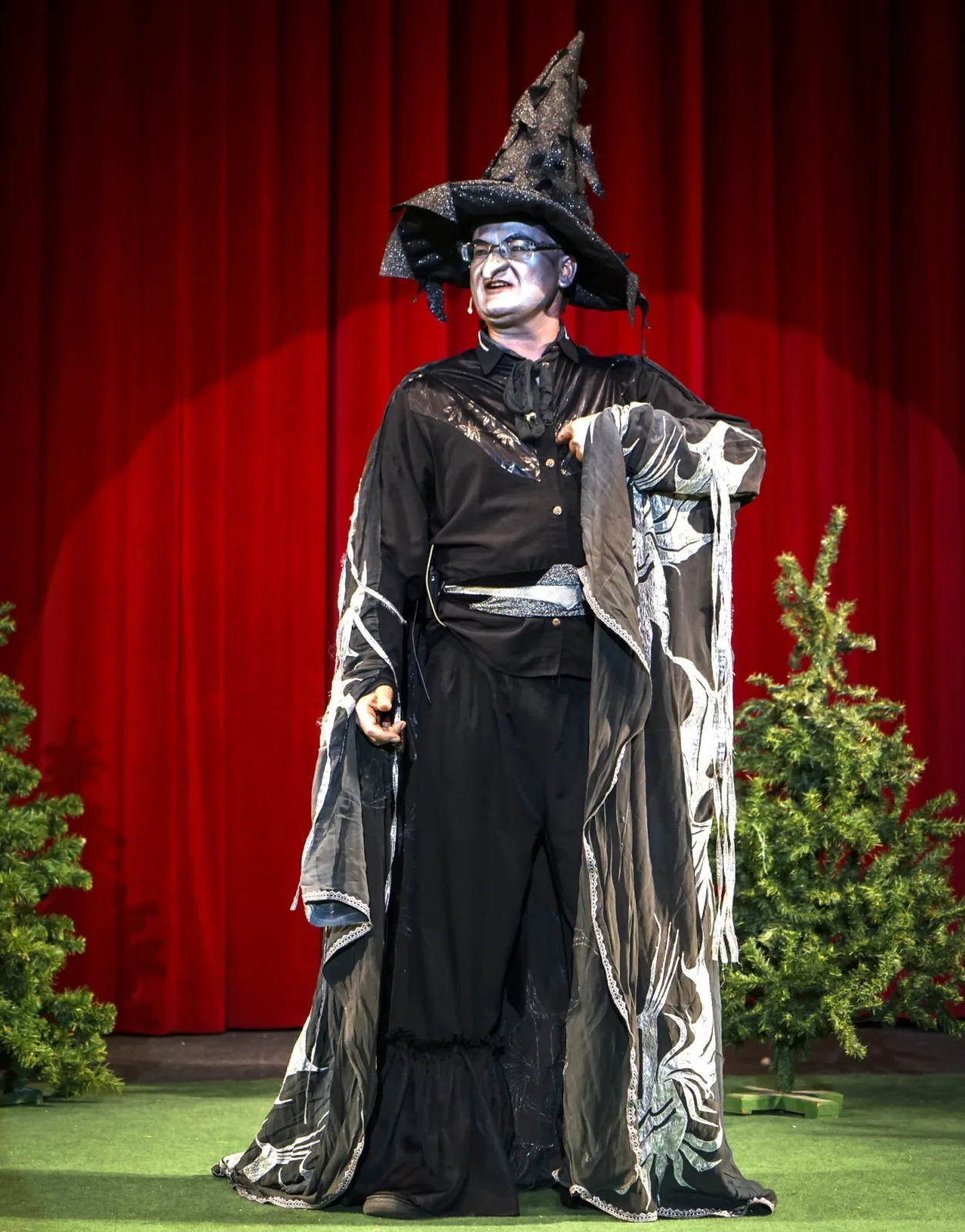 Hexenmeister in schwarzsilbernem Mantel – Ein Zauberer mit funkelndem Spitzhut und silbernem Make up steht vor rotem Vorhang zwischen Tannen und hebt beschwörend die Hand – magischer Auftritt.