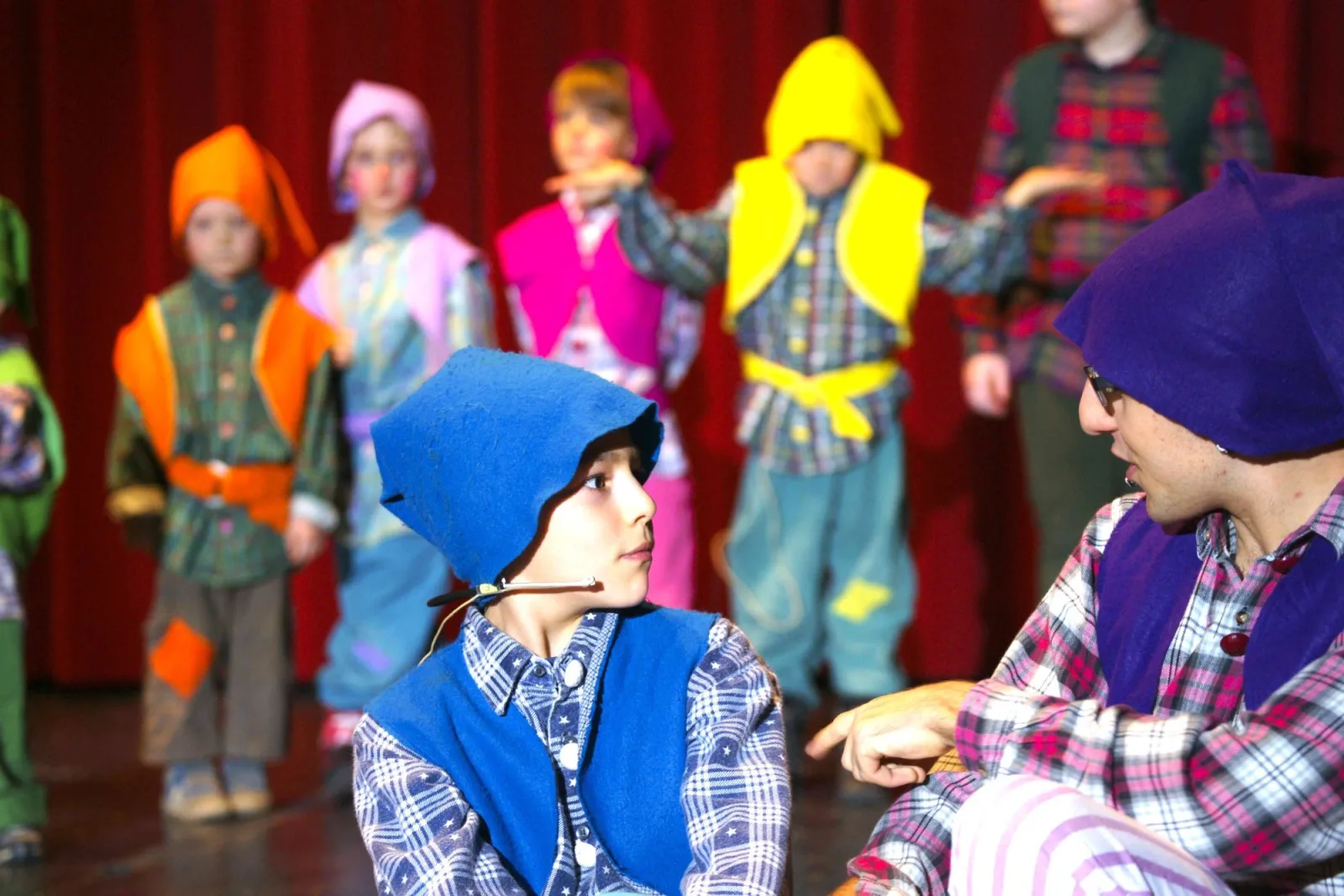 Wichtel im Gespräch auf der Bühne – Ein junger Wichtel mit blauem Häubchen hört aufmerksam einem älteren Wichtel zu, im Hintergrund stehen bunt gekleidete Helfer – Beratung in der Werkstattgeschichte.