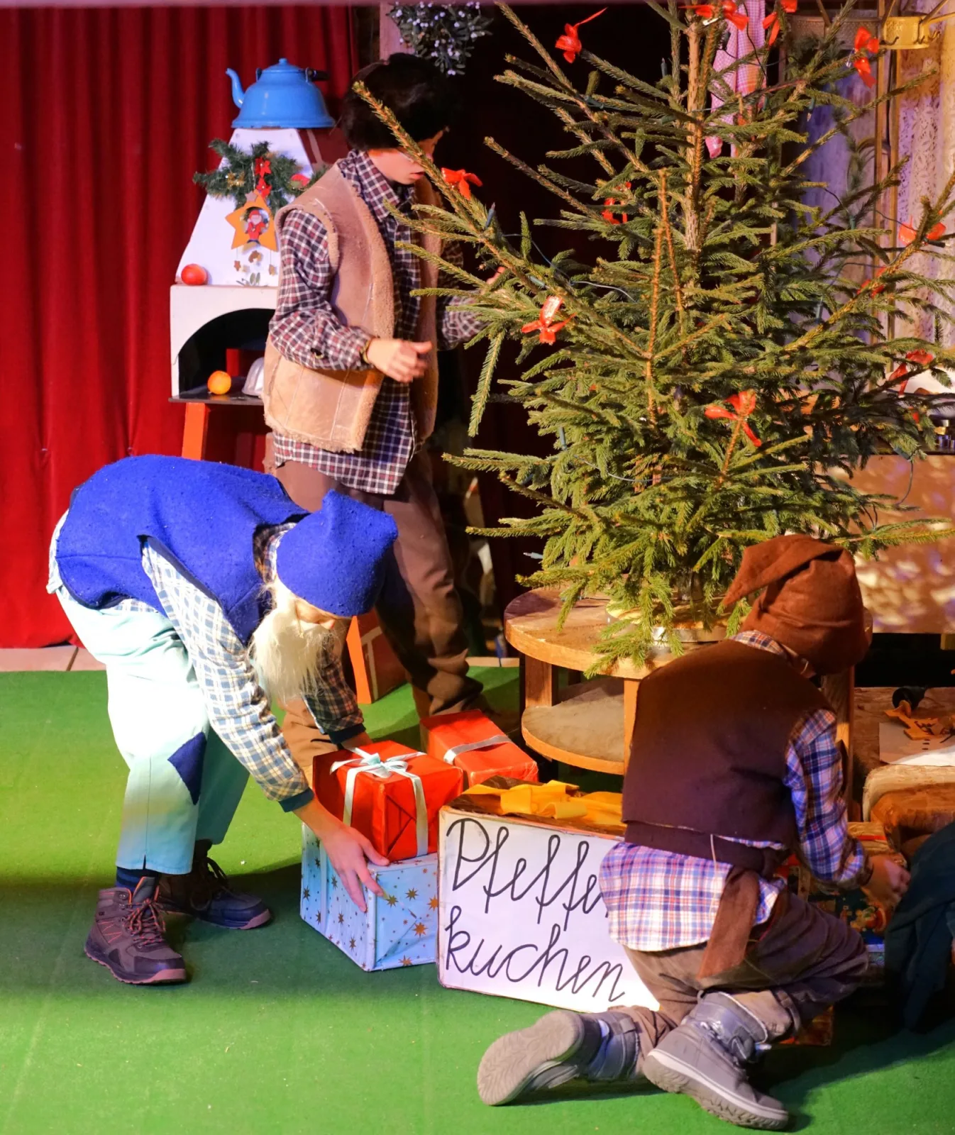 Wichtel legen Geschenke unter den Tannenbaum – Bunt gekleidete Wichtel platzieren große Päckchen neben einer Kiste Pfefferkuchen am Weihnachtsbaum, Ofen und Dekoration sorgen für gemütliche Stubenstimmung.