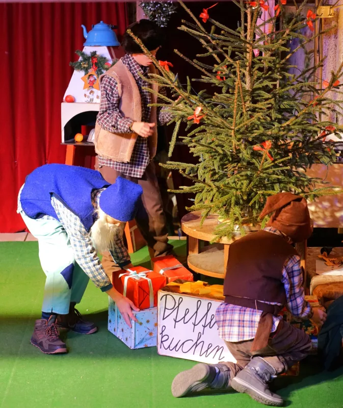 Wichtel legen Geschenke unter den Tannenbaum – Bunt gekleidete Wichtel platzieren große Päckchen neben einer Kiste Pfefferkuchen am Weihnachtsbaum, Ofen und Dekoration sorgen für gemütliche Stubenstimmung.