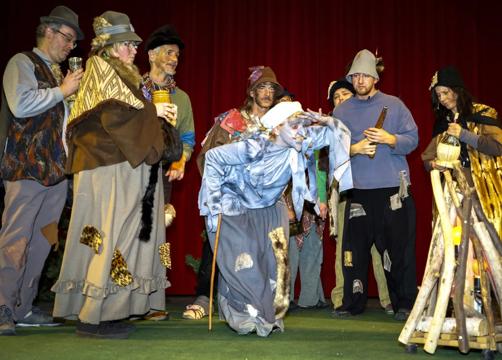 Räuber am Lagerfeuer mit Späherin – Ein Kreis rustikal gekleideter Räuber steht am Holzfeuer, vorne späht die alte Knusperhexe mit Stock in die Ferne – nächtliche Waltszene vor rotem Vorhang.