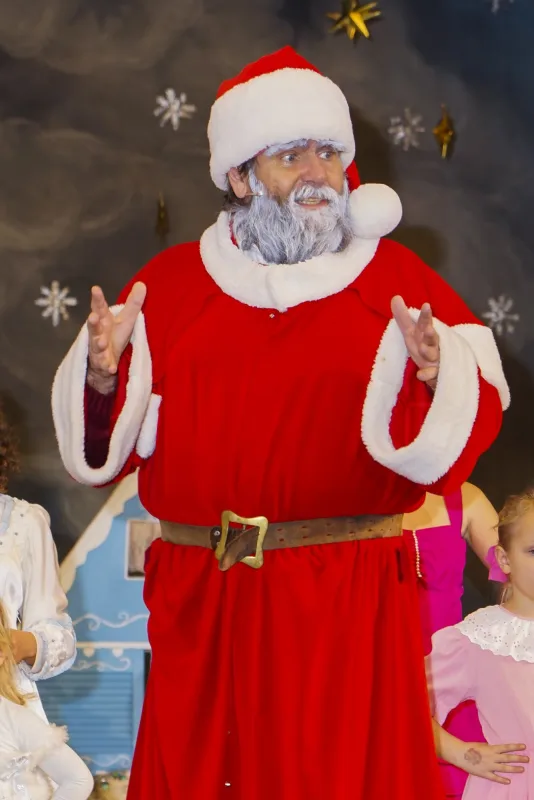 Weihnachtsmann im festlichen Auftritt – Der Weihnachtsmann in roter Robe spricht lächelnd vor winterlichem Hintergrund, Kinder in Engels- und Festtagskleidern stehen neben ihm – herzerwärmender Höhepunkt.