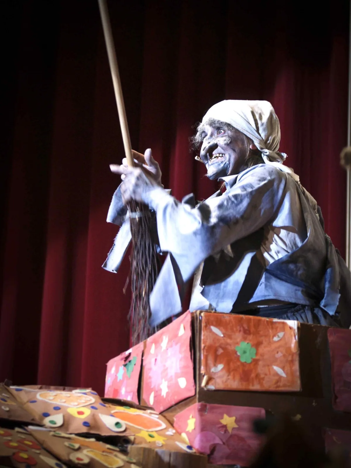 Alte Knusperhexe schwingt den Besen auf dem Knusperhäuschen – Eine grau geschminkte Hexe mit Kopftuch erhebt lachend den Reisigbesen, sie hockt auf dem bunten Lebkuchenhäuschen vor rotem Vorhang – freche Waldmagie mit Humor.