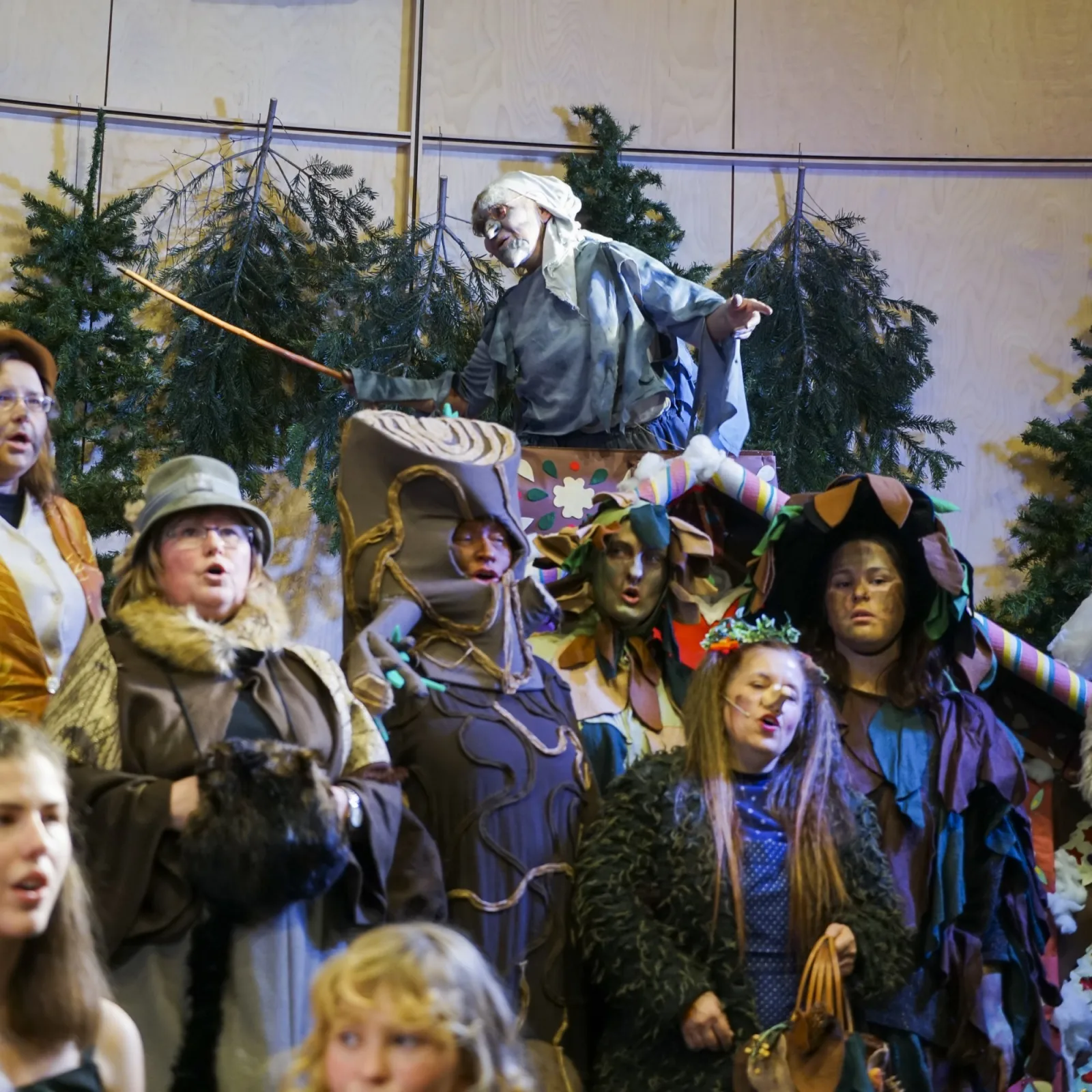 Waldvolk vor dem Knusperhäuschen mit Hexe auf dem Dach – Vor Tannen und dem bunt verzierten Häuschen versammeln sich Baumwesen, Moosgestalten und die Hexe, die vom Dach aus das Geschehen dirigiert – stimmungsvolle Chor-Szene.