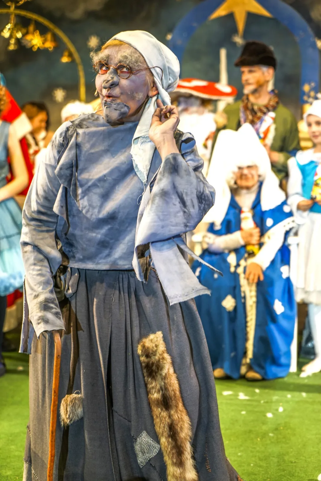 Alte Knusperhexe lauscht vor Sternentor und Himmelsgesellschaft – Die alte Knusperhexe mit Brille und Stock hält die Hand ans Ohr, im Hintergrund schweben Christkind-Gestalten, Sterne und blaue Bögen – komischer Moment im Übergang zum Himmel.