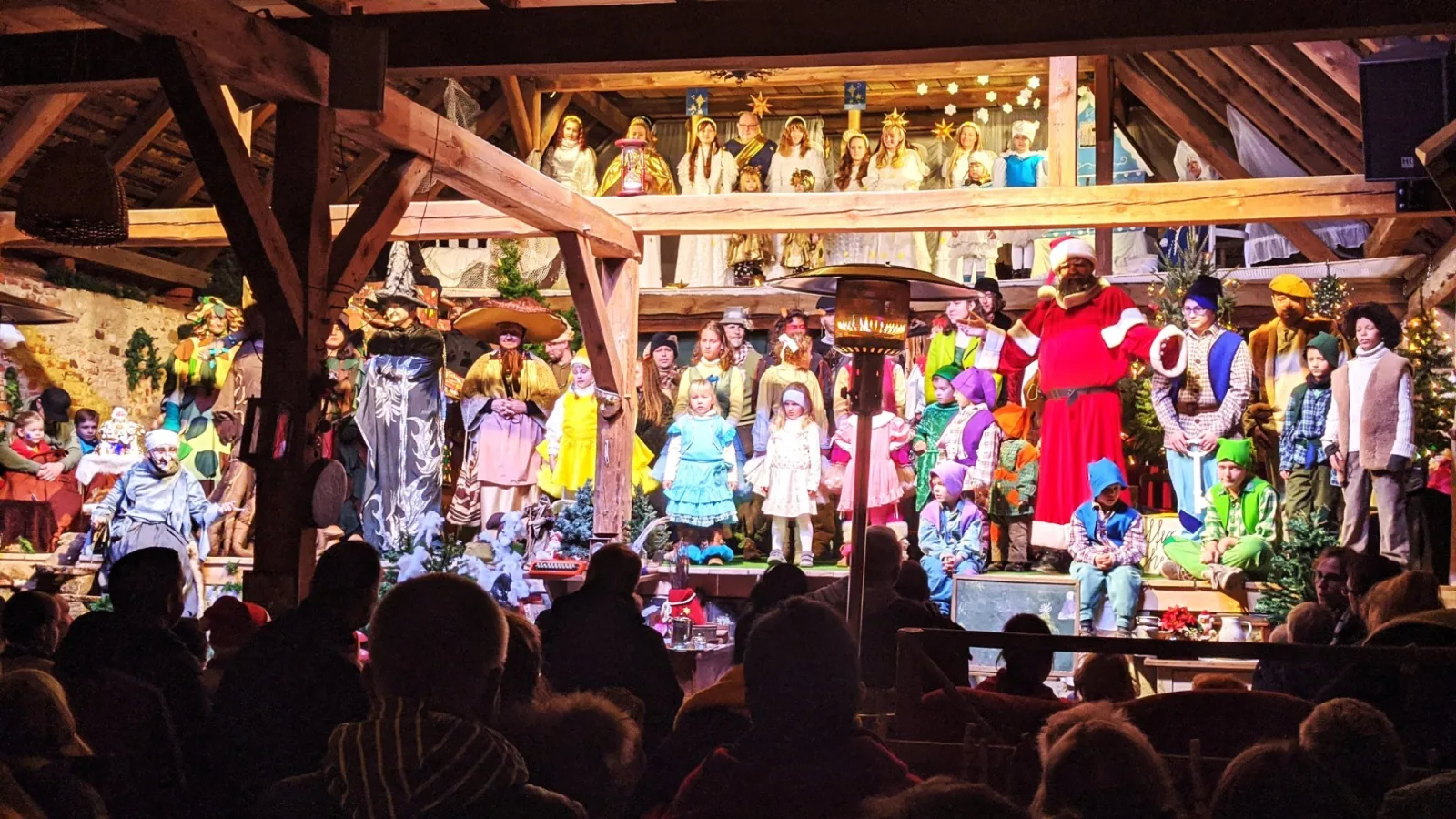 Großes Finale der Aufführung mit Ensemble und Weihnachtsmann – Ein voll besetztes Bühnenbild zeigt Wichtel, Waldwesen, Engel und den Weihnachtsmann, davor staunt das Publikum – festlicher Höhepunkt der Aufführung.