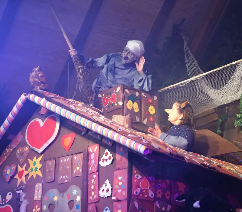 Hexe auf dem Dach des Lebkuchenhauses im Gespräch – Die Hexe lehnt sich mit Besen aus dem Schornstein des Knusperhauses und hält die Hand ans Ohr, unten antwortet die Trude mit langer Nase – schelmisches Streitgespräch.