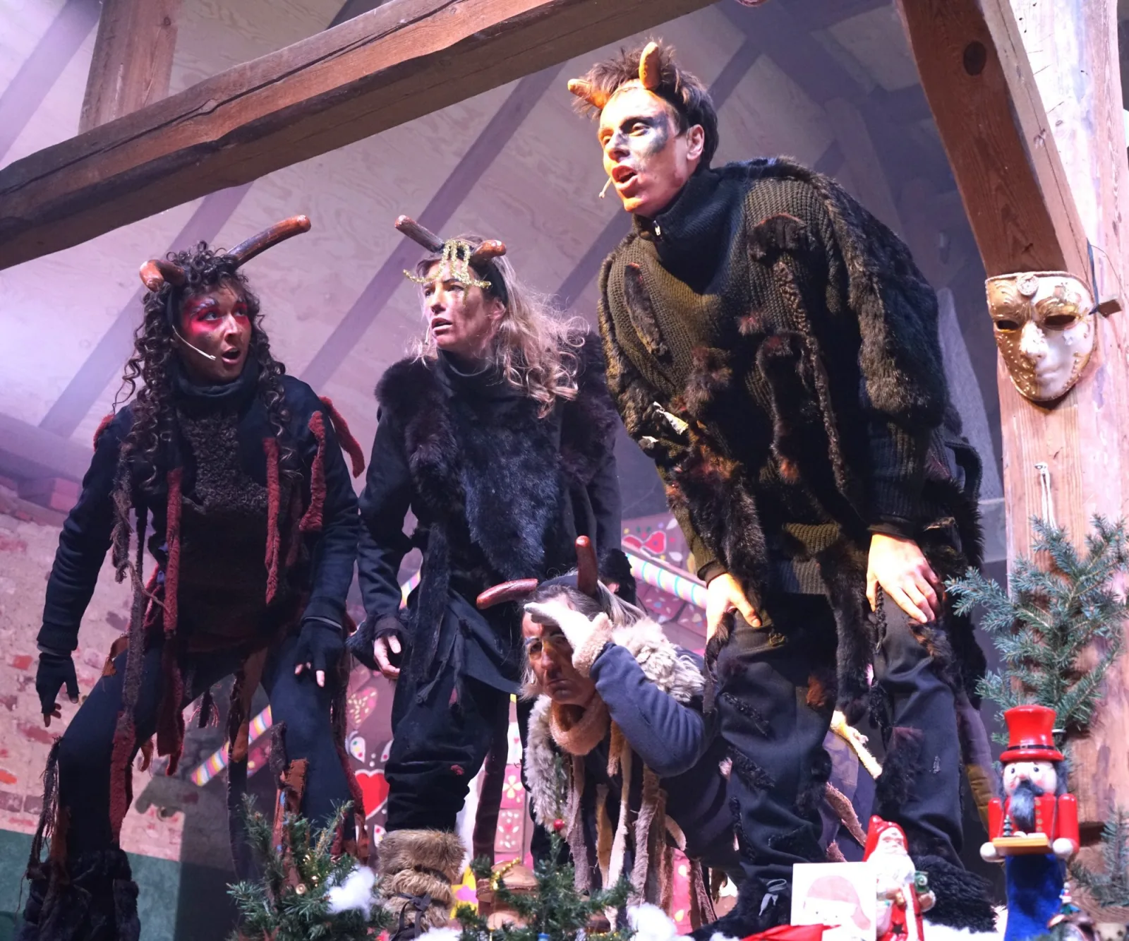 Teufelskinder spielen auf der Märchenbühne – Drei Teufelkinder mit Hörnern und Fell stehen dicht beisammen und rufen in den Raum, neben ihnen weihnachtliche Requisiten – kraftvolles Schauspiel.