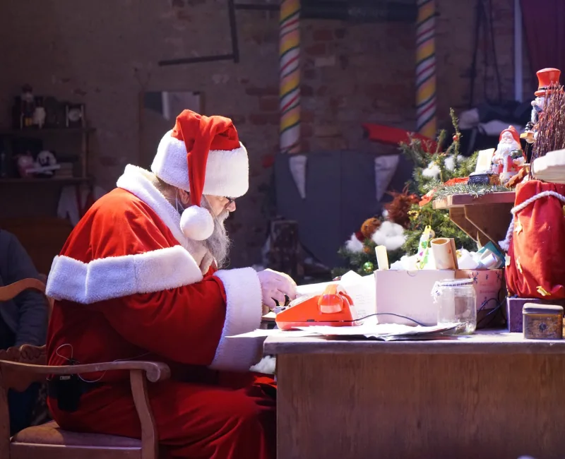 Weihnachtsmann sortiert Wunschbriefe am Schreibtisch – Der Weihnachtsmann arbeitet konzentriert an seinem Holztisch mit rotem Telefon, Listen und kleinen Tannen, Geschenkesäcke stapeln sich – stille Vorbereitung vor der Bescherung.