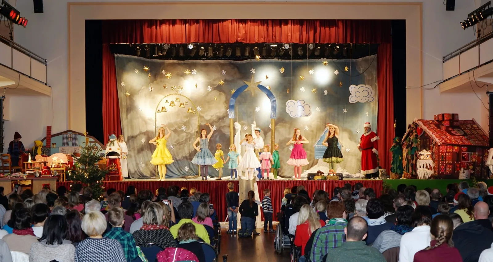 Himmelszelt mit lebendigen Puppen und Weihnachtsmann im Theatersaal – Vor Wolkenhimmel und Sternen tanzen farbige Püppchen unter goldenen Bögen, rechts wartet der Weihnachtsmann, das Publikum füllt den Saal – festliche Bühnenpanorama.