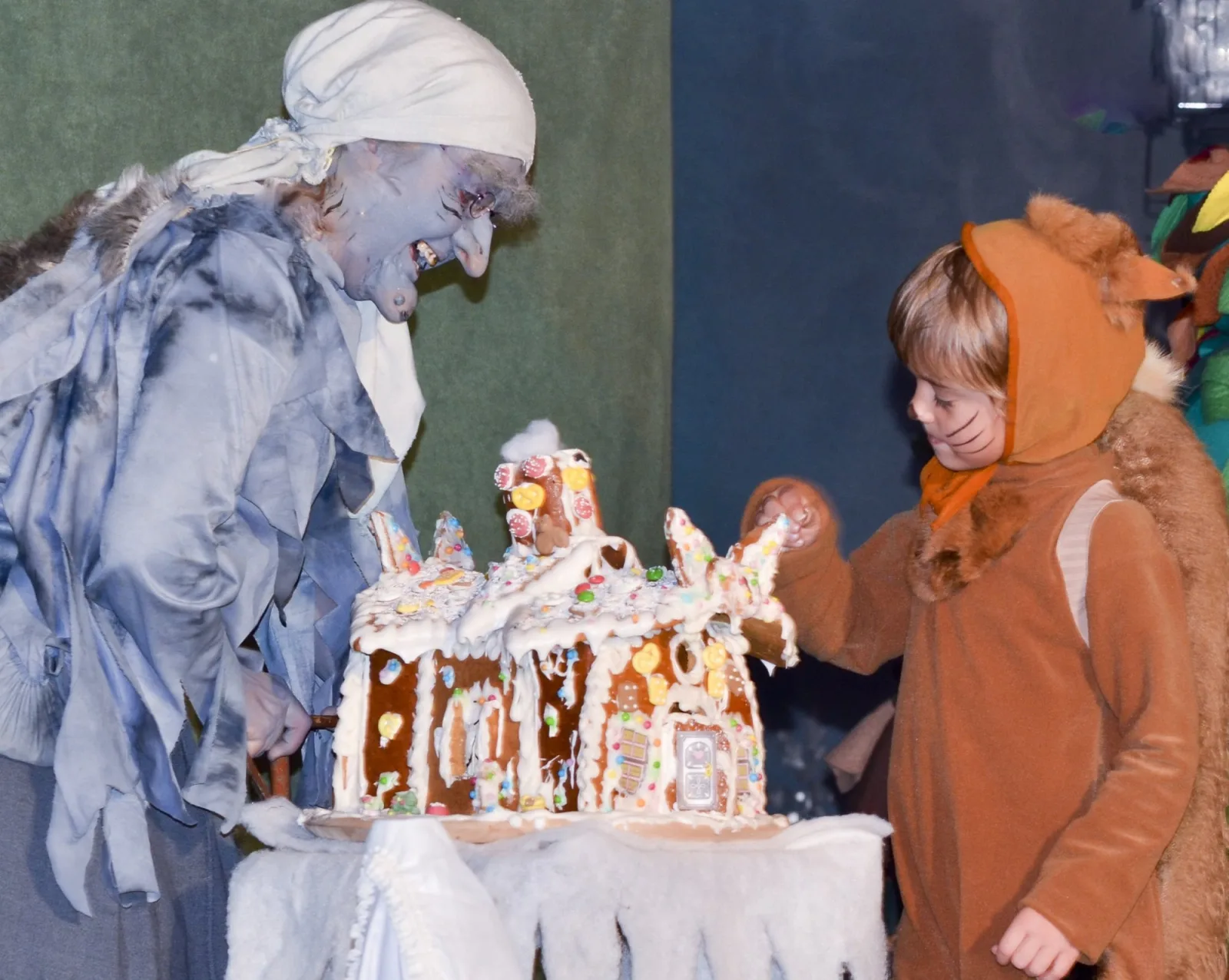 Hexe und Eichhörnchen-Kind am süßen Lebkuchenhaus – Die Hexe grinst zum verkleideten Eichhörnchen-Kind, das neugierig Zuckerguss vom Miniatur-Knusperhaus stibitzt – verspielter Moment zwischen Versuchung und Abenteuer.